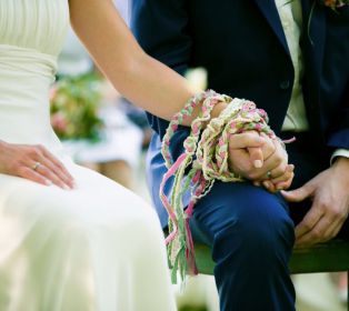 Freie Trauung: Brautpaar sitzend bei der Segnung, rechte Hand Bräutigam und linke Hand Braut umwickelt mit Segensbändern, DIY