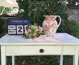 Freie Trauung unter freiem Himmel: alter Wasserkrug bestückt mit Olivenzweigen, nostalgisches Kalenderschild, Brautstrauß in rose-weiß auf Vintage Holztisch mit rosa Spitzen-Leinenläufer, Hochzeitsdeko zu mieten